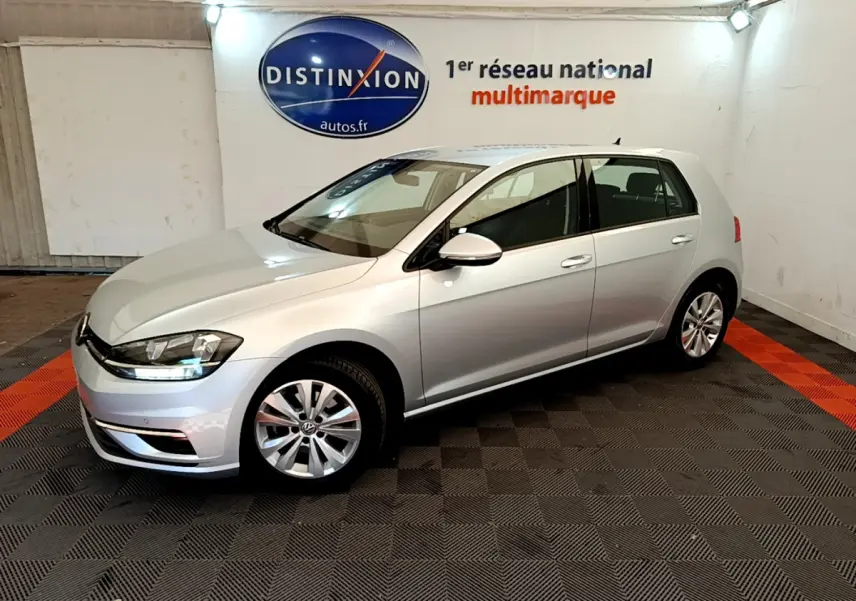 Volkswagen Golf gris clair vue 3/4 avant droit dans un showroom avec logo Distinxion en arrière-plan.