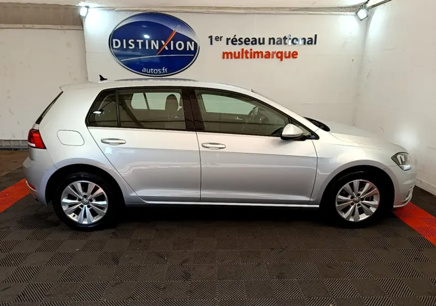 Profil droit d'une Volkswagen Golf gris clair 2019 garée en intérieur avec logo Distinxion en arrière-plan.