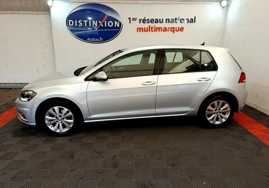Profil droit d'une Volkswagen Golf gris clair 2019 garée en intérieur sur sol à damiers noir et orange.