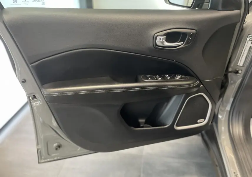 Porte avant gauche ouverte du Jeep Compass gris foncé, montrant garniture noire avec surpiqûres blanches et haut-parleur intégré.
