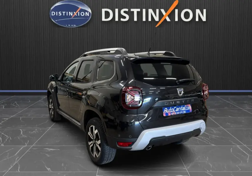 Vue 3/4 arrière droite d'un Dacia Duster noir 2022 avec jantes bi-ton et barres de toit visibles en intérieur showroom.
