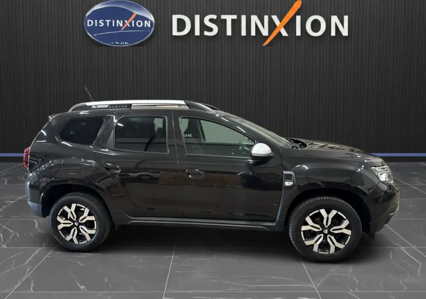 Vue de profil côté gauche d'un Dacia Duster noir 2022 avec jantes bi-ton et rétroviseurs blancs, sur fond intérieur showroom.