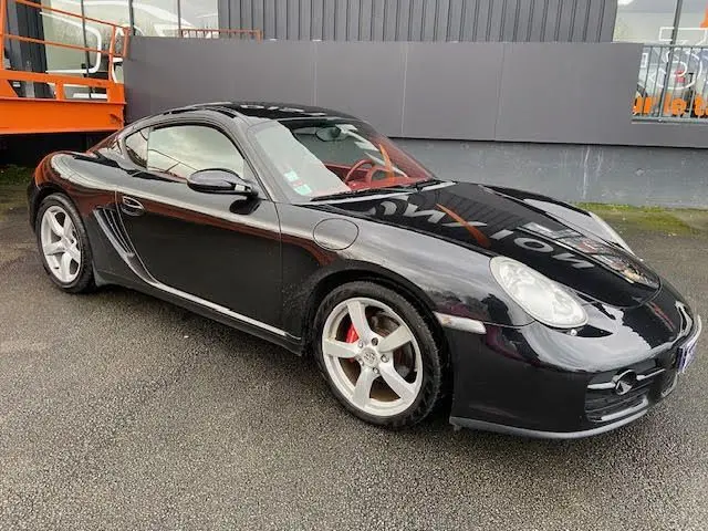 Porsche Cayman noir vue 3/4 avant droit, jantes argentées et intérieur cuir rouge visible au travers des vitres.