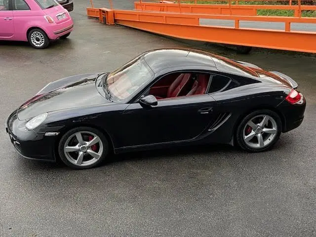 Vue de profil côté gauche d'une Porsche Cayman noire de 2008 avec intérieur cuir rouge et jantes alliage cinq branches.