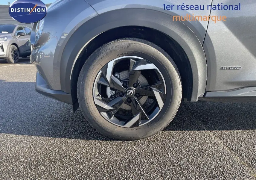 Gros plan sur la roue avant gauche du Nissan Juke gris squale métal 2024 avec jante alliage bicolore et badge Hybrid.