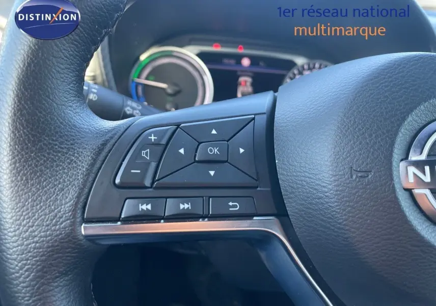 Gros plan sur les commandes au volant d'un Nissan Juke 2024 gris squale métal, avec tableau de bord hybride en arrière-plan.