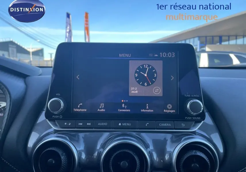 Écran tactile central du tableau de bord du Nissan Juke gris, affichant l'horloge et les options multimédia.