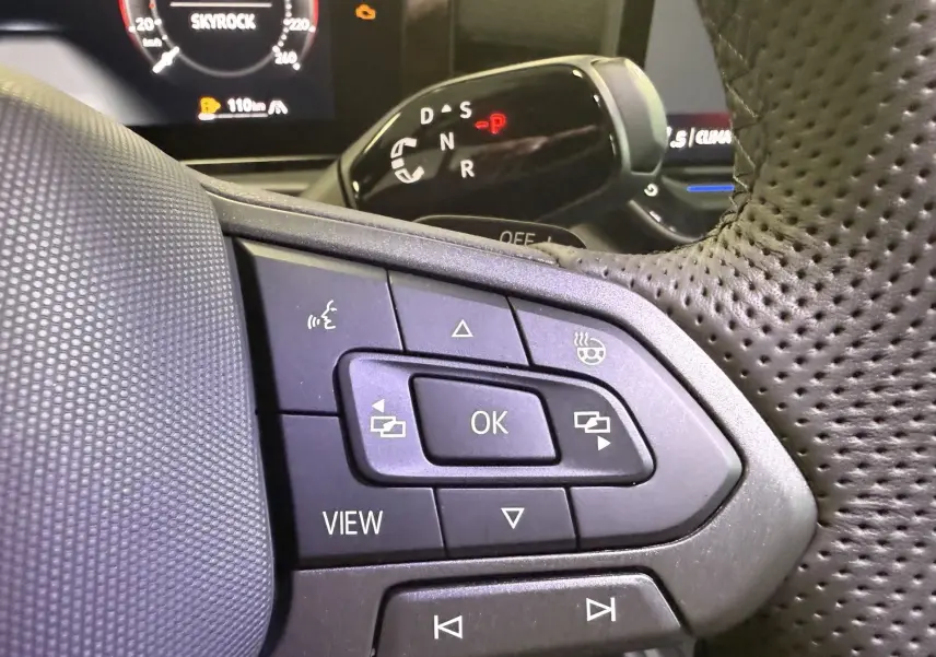 Gros plan sur les commandes multifonctions à gauche du volant perforé du Volkswagen T-Roc noir intense, avec affichage digital en arrière-plan.