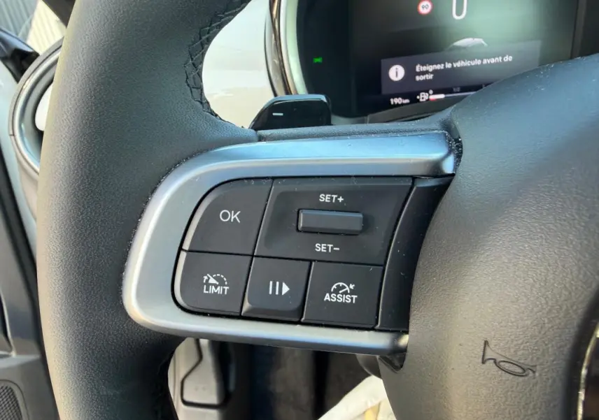 Gros plan sur les commandes du régulateur de vitesse au volant de la FIAT 600 blanc, tableau de bord numérique en arrière-plan.