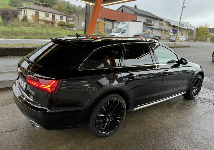 Audi A6 Avant Allroad noire vue 3/4 arrière droit, avec jantes noires et feux arrière allumés sous abri.