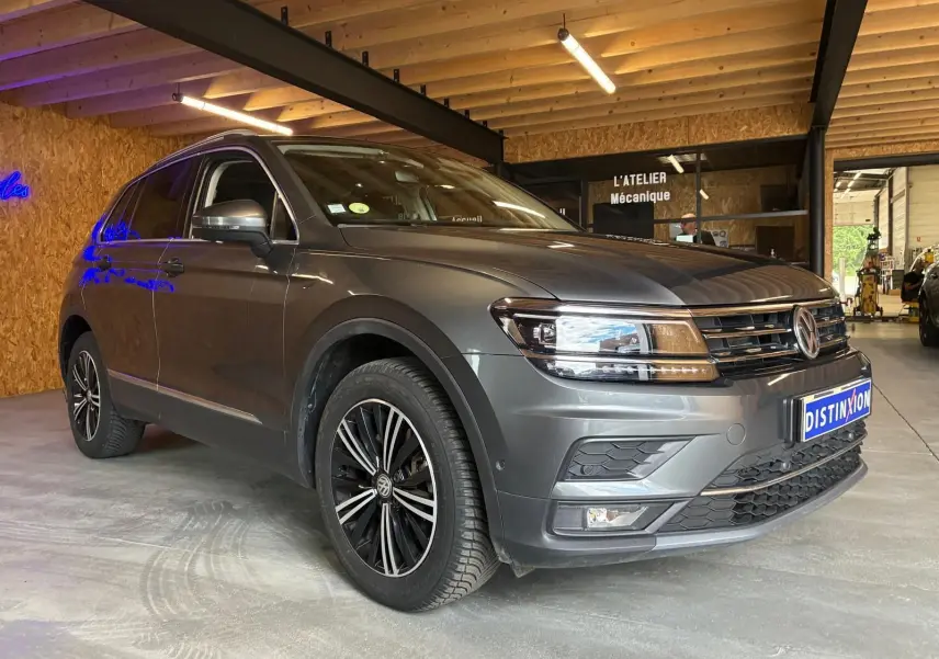 Volkswagen Tiguan gris 3/4 avant droit, avec jantes noires et détails chromés, stationné en intérieur d'atelier.