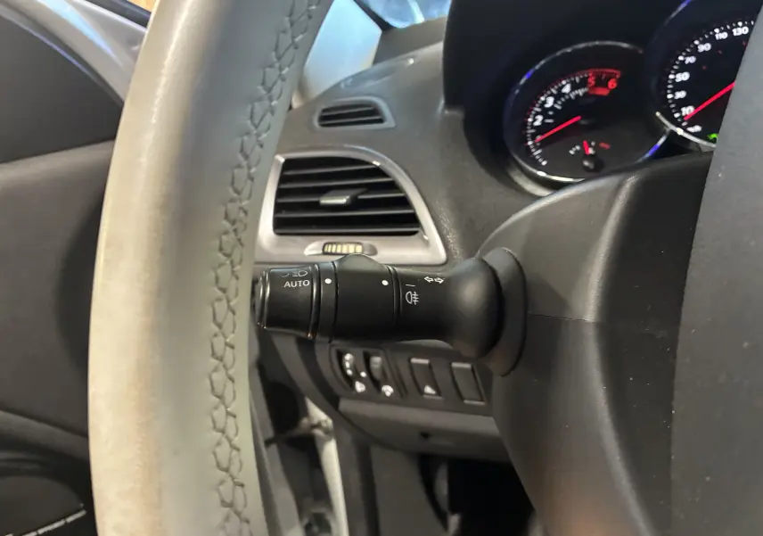 Gros plan sur le volant cuir et la commande des phares automatiques à gauche du tableau de bord d'une Renault Mégane Coupé blanche.