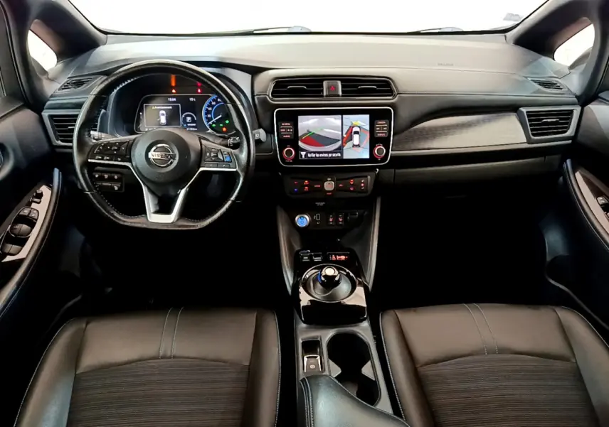Vue intérieure avant du Nissan Leaf blanc 2020, volant cuir, écran tactile central affichant la caméra 360° d'aide au stationnement.