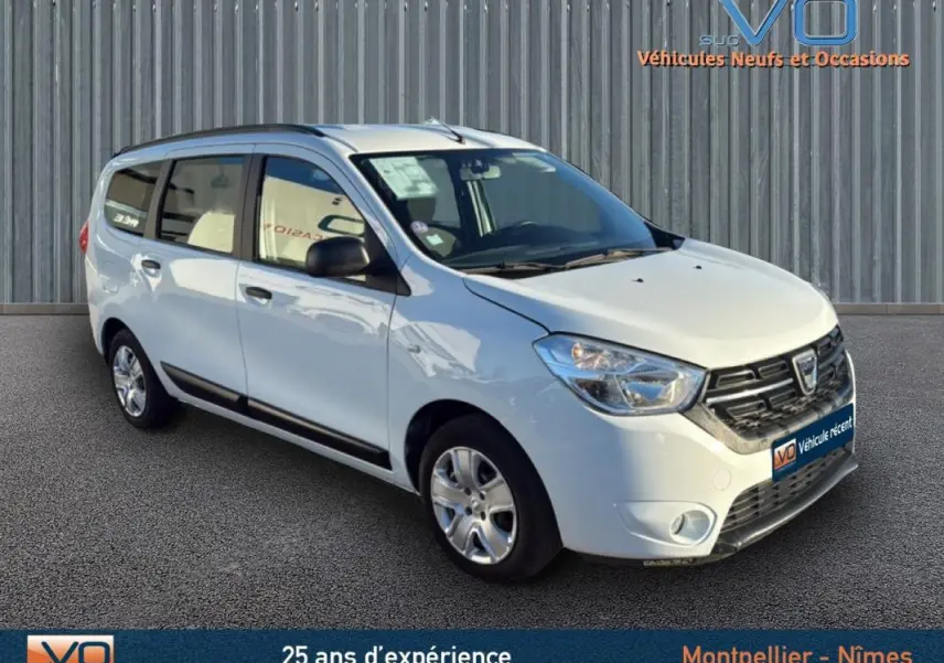Vue 3/4 avant d'un Dacia Lodgy blanc avec barres de toit, garé devant un mur métallique.