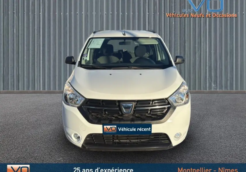 Vue frontale d'un Dacia Lodgy blanc avec calandre noire et barres de toit, stationné devant un mur métallique.