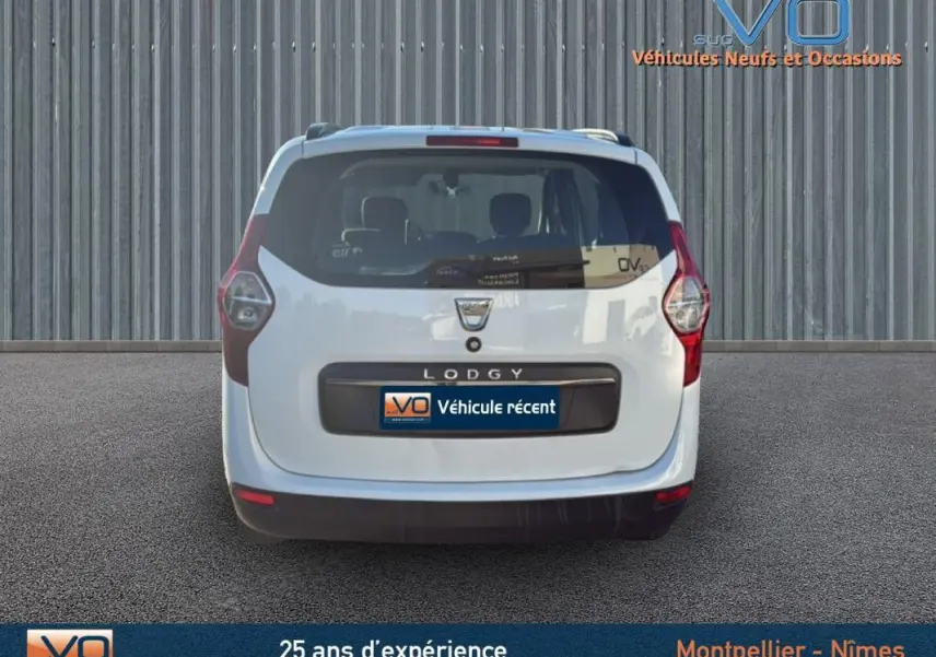 Vue arrière d'un Dacia Lodgy blanc avec barres de toit, sur fond industriel gris.