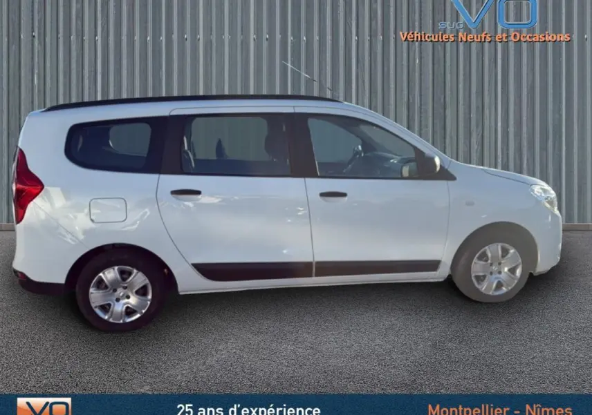 Profil droit du Dacia Lodgy blanc 2018 avec barres de toit, vitres teintées et jantes acier en arrière-plan urbain.