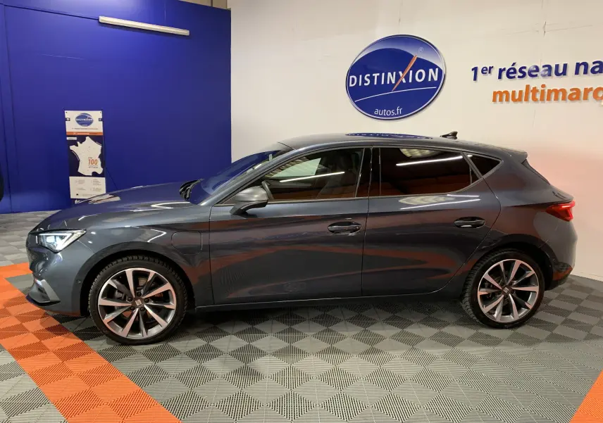 Profil côté gauche d'une SEAT LEON E-HYBRID 204 FR gris métallisé, avec jantes alliage et vitres teintées, en intérieur showroom.
