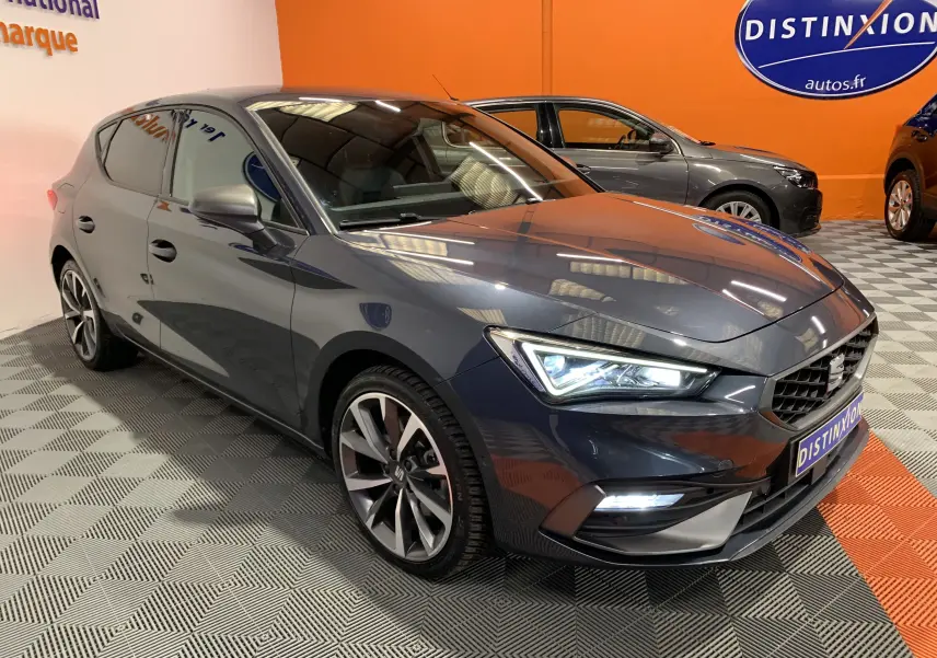 Vue 3/4 avant droite d'une SEAT Leon E-HYBRID gris métallisé avec phares LED allumés en intérieur showroom.