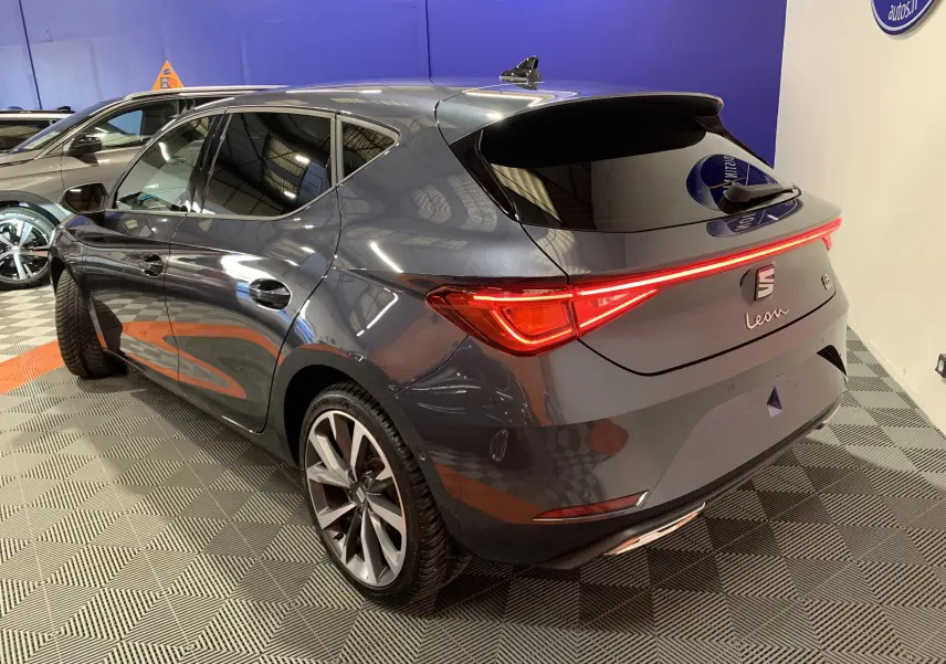 Vue 3/4 arrière droite d'une SEAT Leon E-HYBRID 204 FR gris métallisé avec feux arrière LED allumés en intérieur showroom.