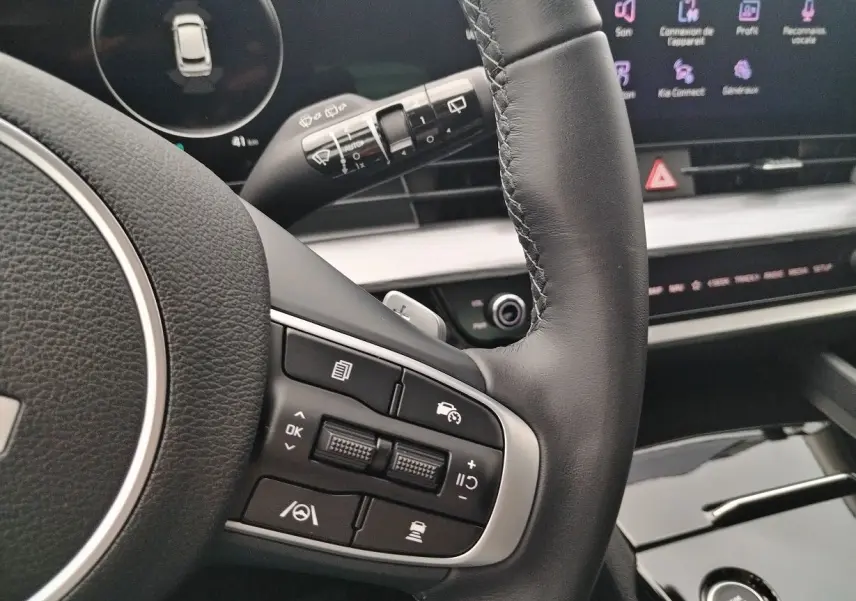 Détail du volant en cuir noir du KIA Sportage 2025, avec commandes intégrées et tableau de bord numérique en arrière-plan.