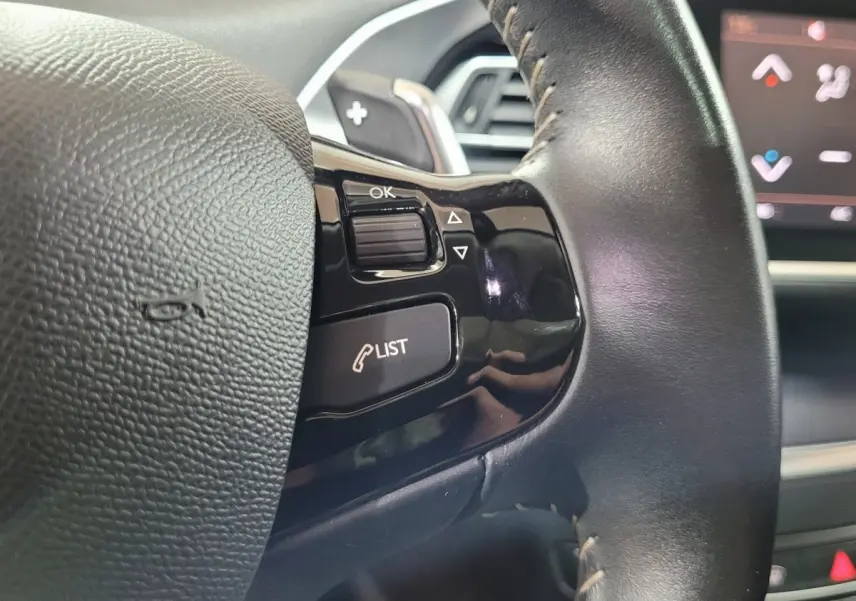 Gros plan sur le volant cuir noir de la Peugeot 308 gris foncé, avec commandes intégrées et écran tactile en arrière-plan.