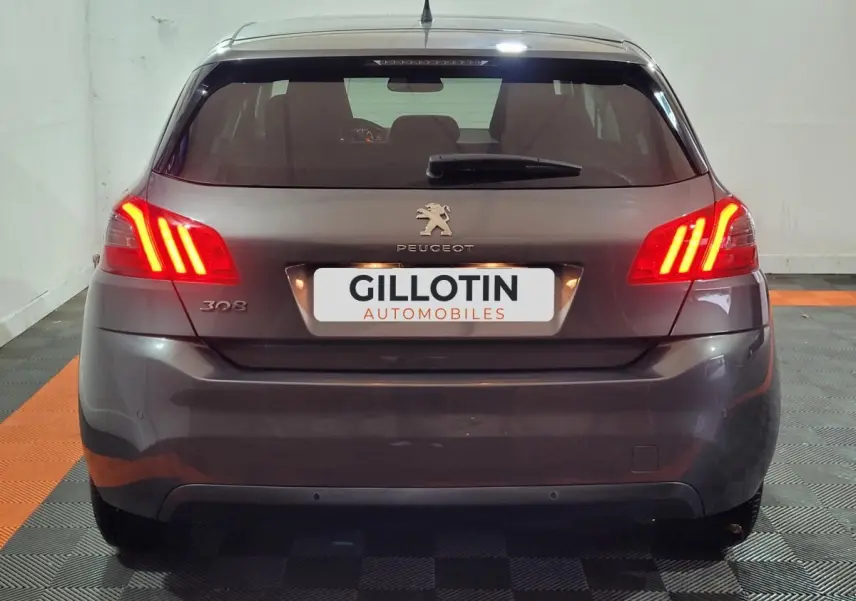 Vue arrière d'une Peugeot 308 gris foncé avec feux arrière allumés et logo visible sur un sol damier noir et orange.