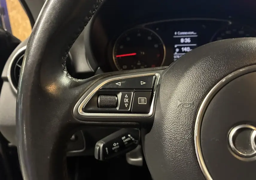 Gros plan sur le volant noir de l'Audi A1 2016, montrant les commandes multifonctions côté gauche et le tableau de bord en arrière-plan.