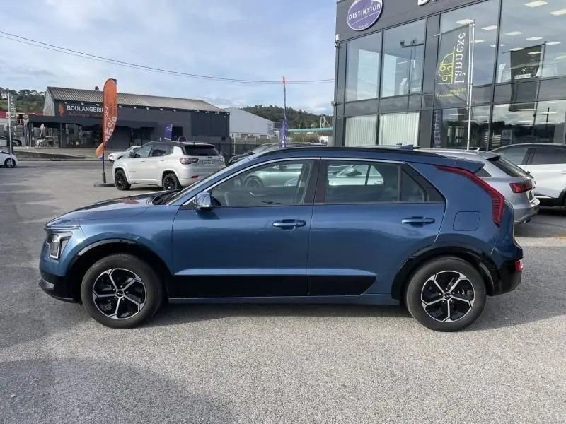 Vue de profil côté gauche d'un KIA NIRO 2024 bleu métal, hybride essence avec jantes noires et détails noirs sur les bas de caisse.