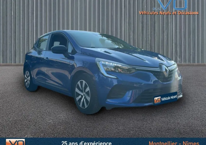 Renault Clio V TCe 90 Equilibre bleu foncé vue 3/4 avant droit sur sol bitumé devant un mur métallique.