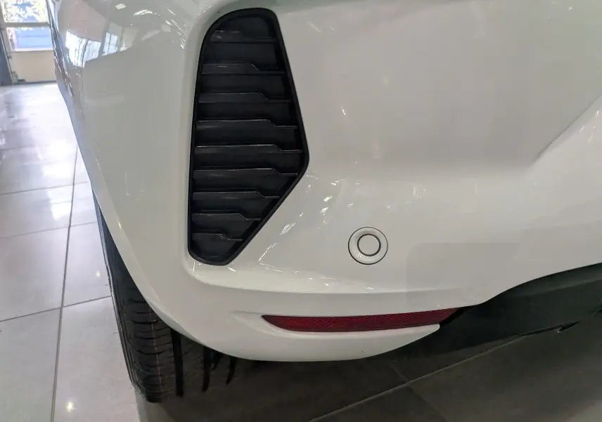 Gros plan sur le pare-chocs arrière gauche blanc glacier de la Renault Clio V avec capteur de recul et prise d'air noire.