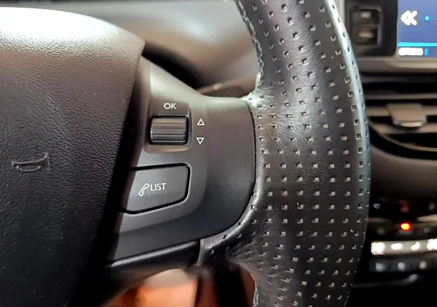 Gros plan sur les commandes gauche du volant en cuir perforé du Peugeot 2008 gris foncé, avec écran tactile en arrière-plan.