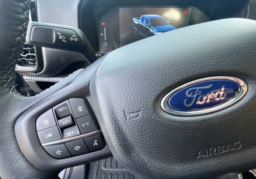 Gros plan sur le volant cuir multifonctions d’un Ford Ranger 2025 avec écran digital affichant un pick-up bleu.