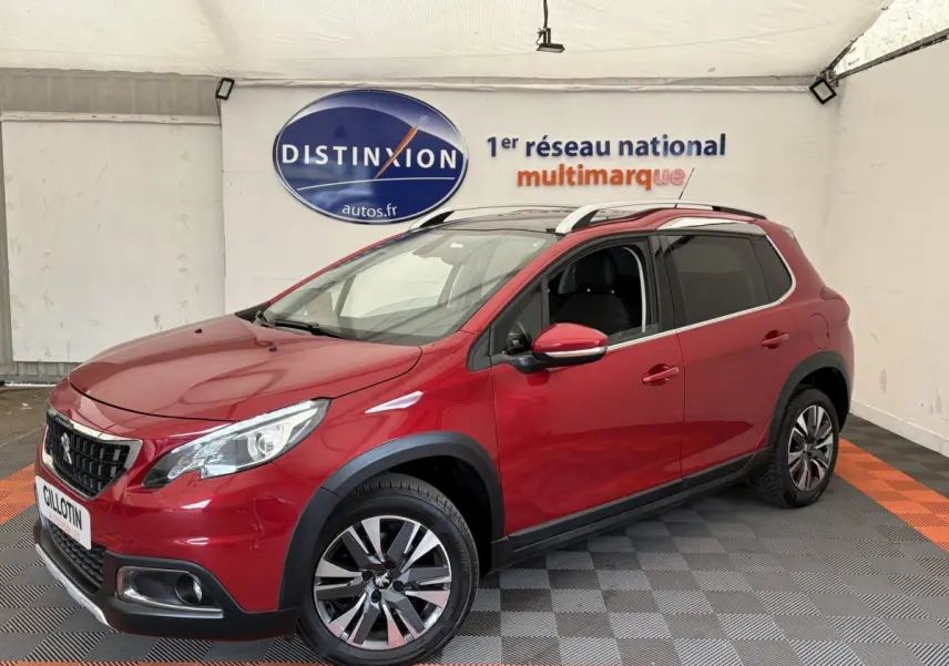 Peugeot 2008 rouge vue 3/4 avant droit, avec jantes alliage et barres de toit chromées en intérieur showroom.