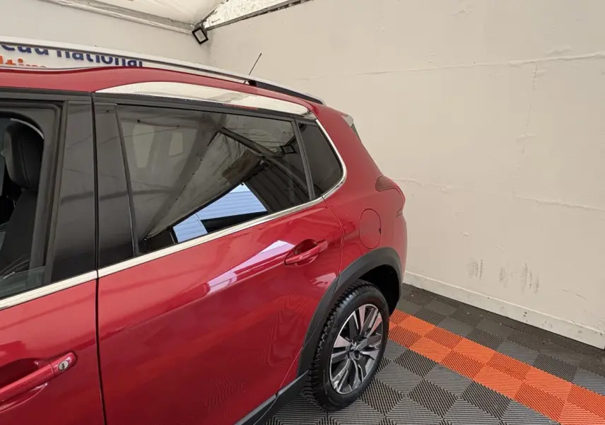 Vue côté droit arrière du Peugeot 2008 rouge avec jantes alliage et barres de toit chromées dans un garage.