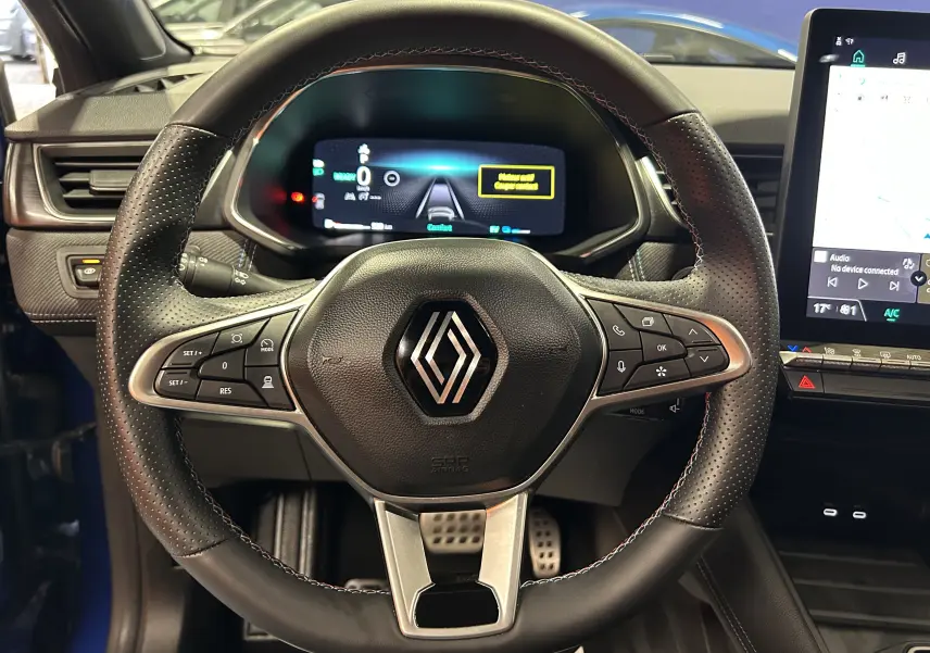 Vue rapprochée du volant noir perforé avec logo Renault, tableau de bord numérique et écran tactile du Renault Captur bleu Iron.