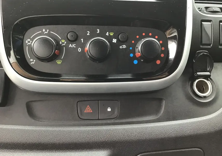 Gros plan sur les commandes de climatisation noires du Renault Trafic blanc, avec bouton d'alerte et prise allume-cigare visible.