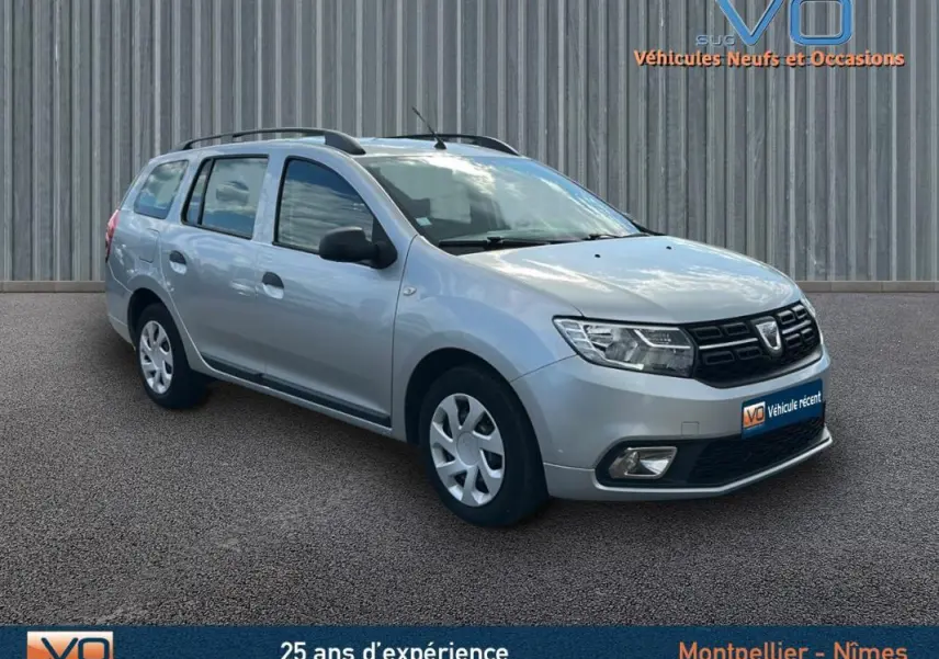 Vue 3/4 avant droite d'une Dacia Logan MCV gris clair avec barres de toit et jantes 15 pouces, stationnée sur sol bitumé.
