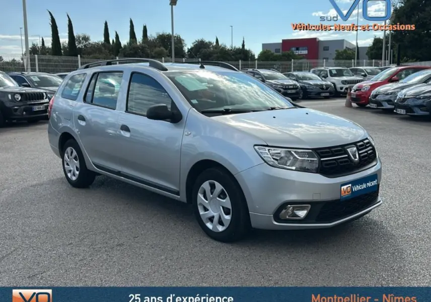 Vue 3/4 avant gauche d'une Dacia Logan MCV gris clair avec barres de toit, stationnée en extérieur sur un parking.