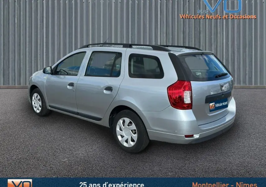 Vue 3/4 arrière droite d'une Dacia Logan MCV gris clair avec barres de toit et jantes 15 pouces en environnement urbain.