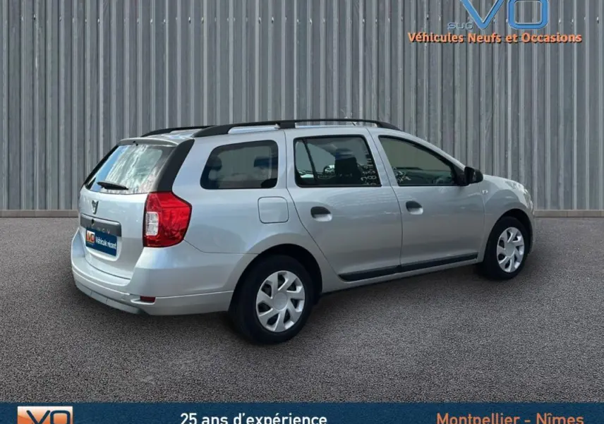 Vue 3/4 arrière droite d'une Dacia Logan MCV gris clair avec barres de toit, sur fond neutre industriel.