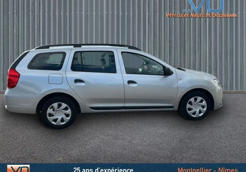 Profil côté droit d'une Dacia Logan MCV gris clair avec barres de toit, garée devant un mur métallique.
