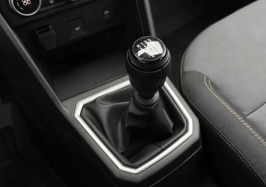 Gros plan sur le levier de vitesses manuel noir entouré d’un cadre argenté, siège avant gris avec surpiqûres beige visible.