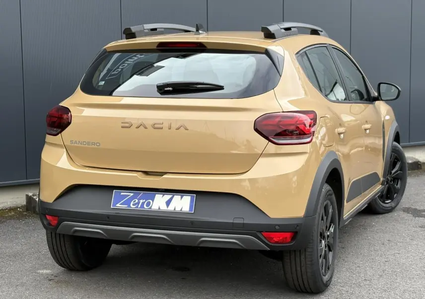 Vue 3/4 arrière droite d'une Dacia Sandero Stepway beige Safari avec barres de toit et jantes noires.