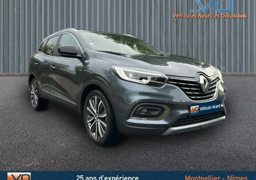 Renault Kadjar gris 3/4 avant droit avec jantes alliage 18 pouces et barres de toit visibles sur sol asphalté.