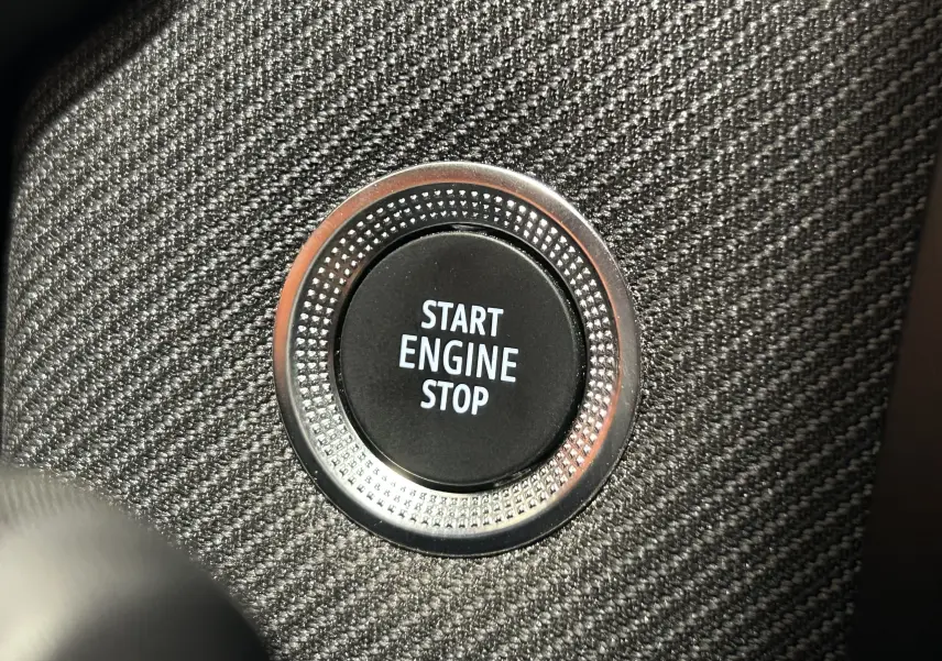 Gros plan sur le bouton Start Engine Stop entouré d’un cerclage métallique, sur fond de tissu gris texturé intérieur Renault Clio V.
