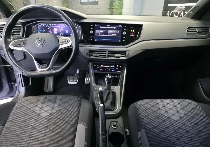 Vue intérieure avant du Volkswagen Taigo 1.5 TSI R-Line, volant cuir multifonctions, tableau de bord digital et sièges tissu noirs à motifs.