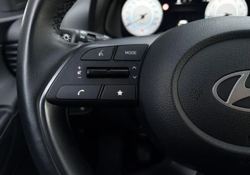 Gros plan sur les commandes gauche du volant cuir noir de la Hyundai i20 2025 avec compteurs numériques en arrière-plan flou.