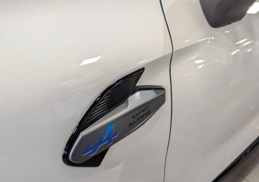 Détail du logo Esprit Alpine bleu sur l'aile avant gauche d'une Renault Symbioz blanc nacré.