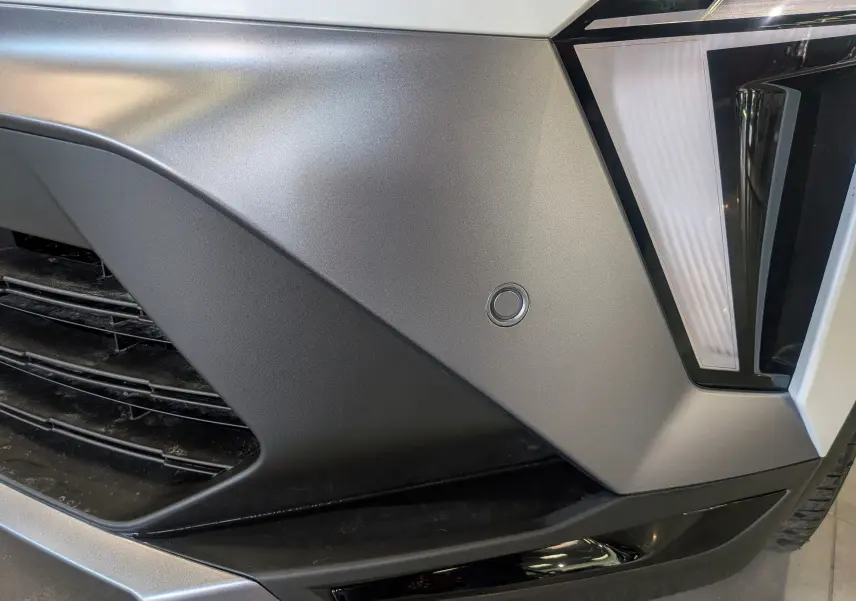 Gros plan sur le pare-chocs avant droit gris mat du Renault Symbioz blanc nacré, avec capteur de stationnement circulaire visible.